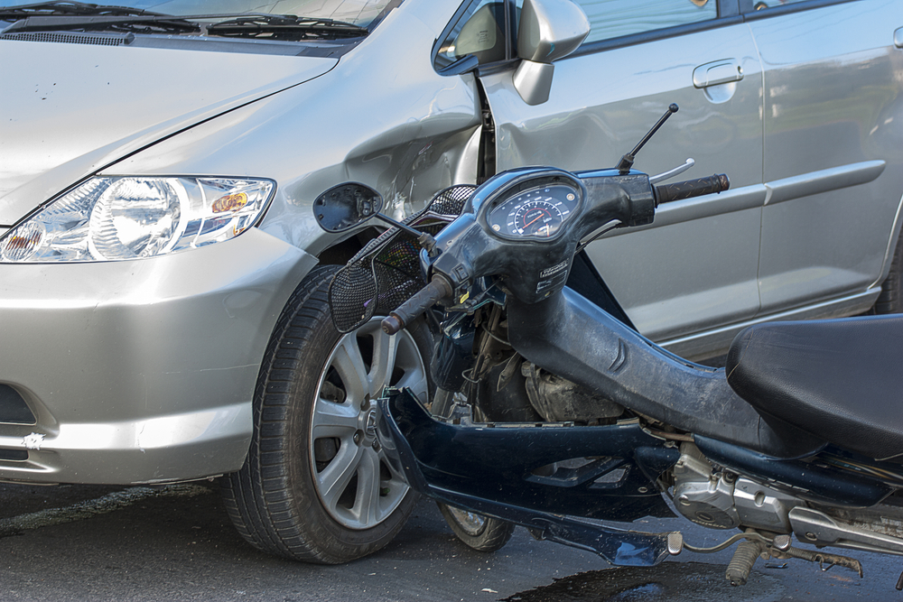 A damaged motorcycle leaning against a silver car after a collision on a Brooklyn street.